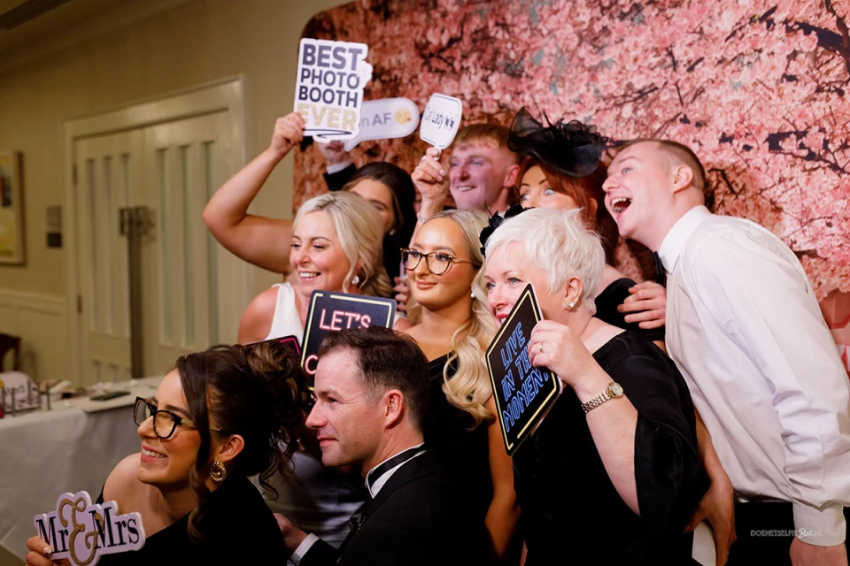 Groep vrienden poseert lachend voor een roze bloemenmuur met een photobooth foto in handen.