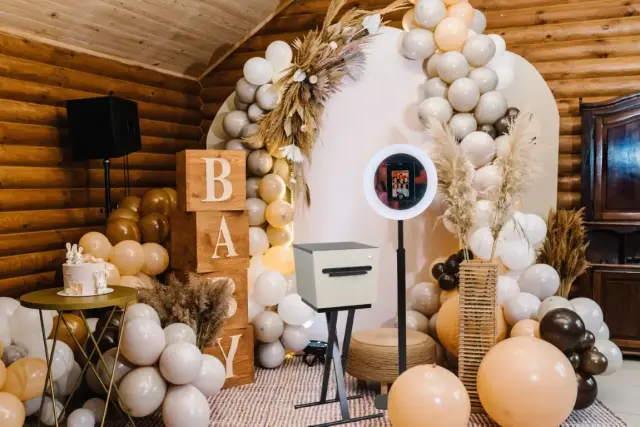 Sfeervol ingerichte babyshower met neutraal gekleurde ballonnen, houten blokken met “BABY”, droogbloemen en een photobooth met ringlamp voor gasten. Alles staat tegen een ronde backdrop in een houten ruimte.