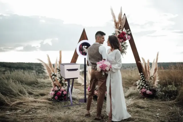 Bruidspaar poseert bij een boho trouwaltaar met pampasgras, bloemen en een moderne photobooth in een open veld.