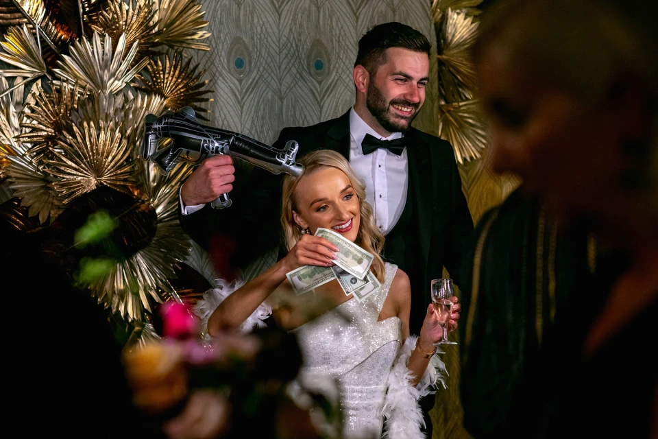 Stijlvolle gasten poseren voor een glitterwand met gouden accenten en een witte photobooth op een galafeest.