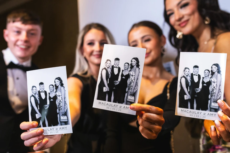 Vriendengroep houdt zwart-wit photobooth-strookjes omhoog met grote glimlach op een jubileumfeest.