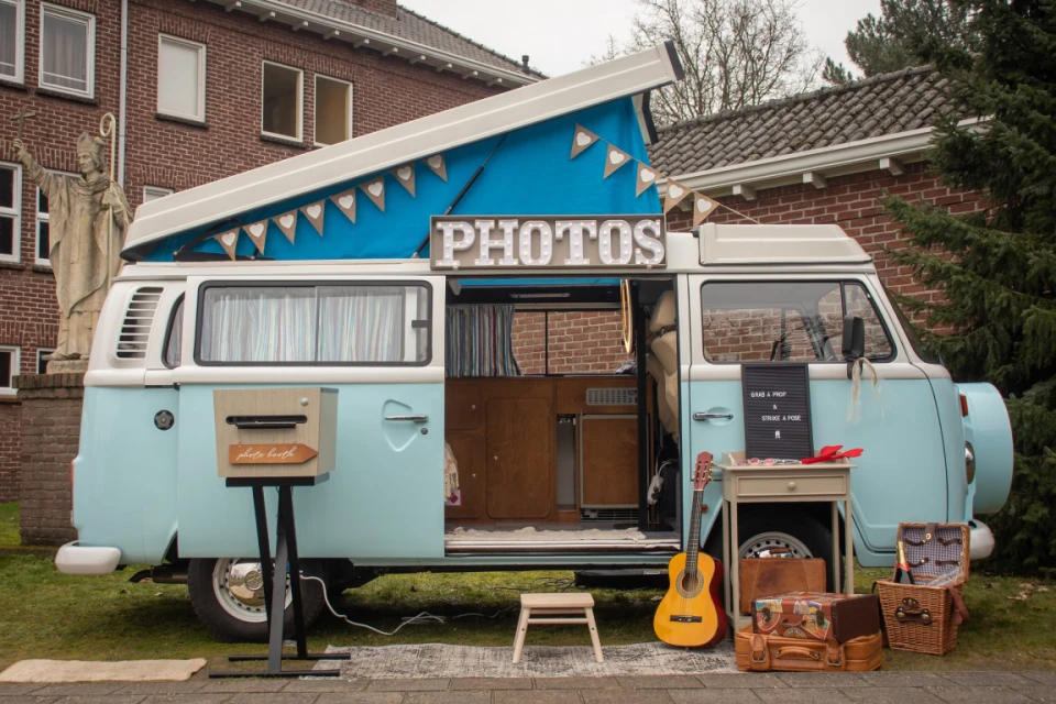 Blauwe VW-bus omgebouwd tot photobooth met &ldquo;PHOTOS&rdquo; bord, aangekleed met koffer, gitaar en retrodetails op een grasveld.